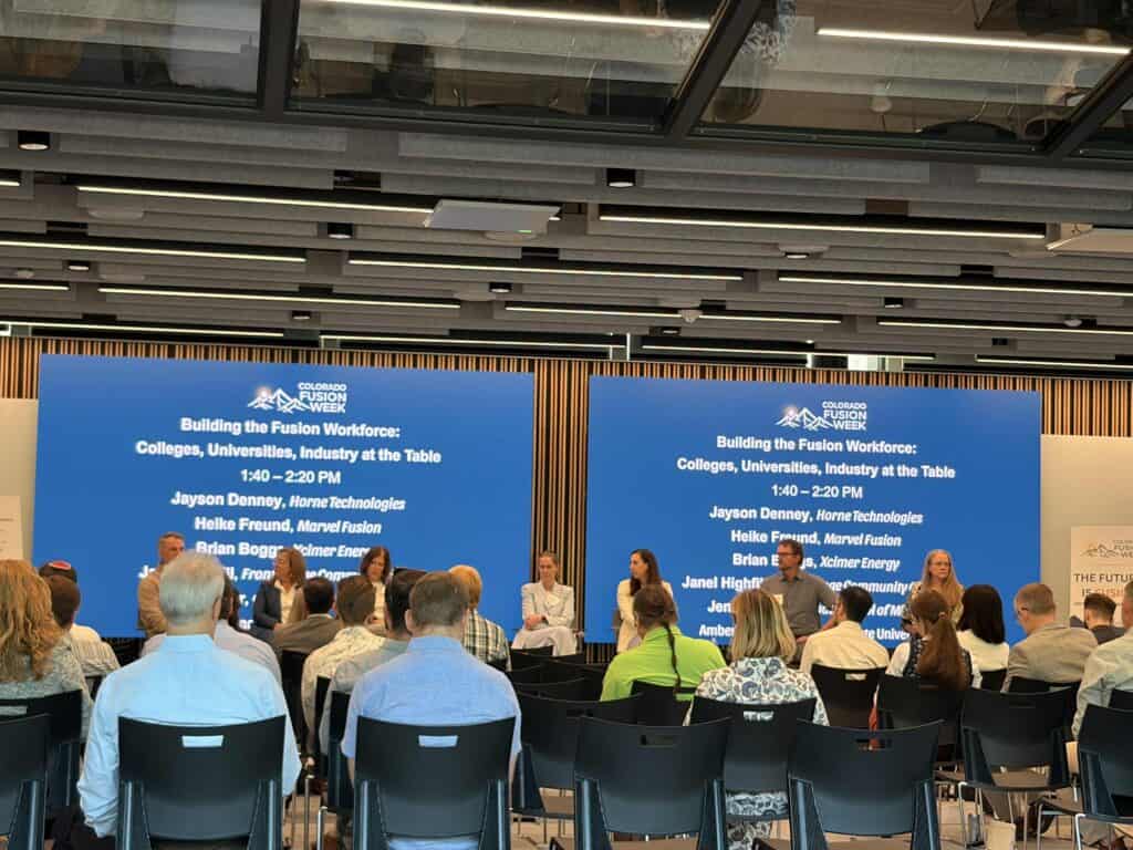 Conference stage with blue panels and event branding reading 'Colorado Fusion Week'; audience seated facing the panelists.